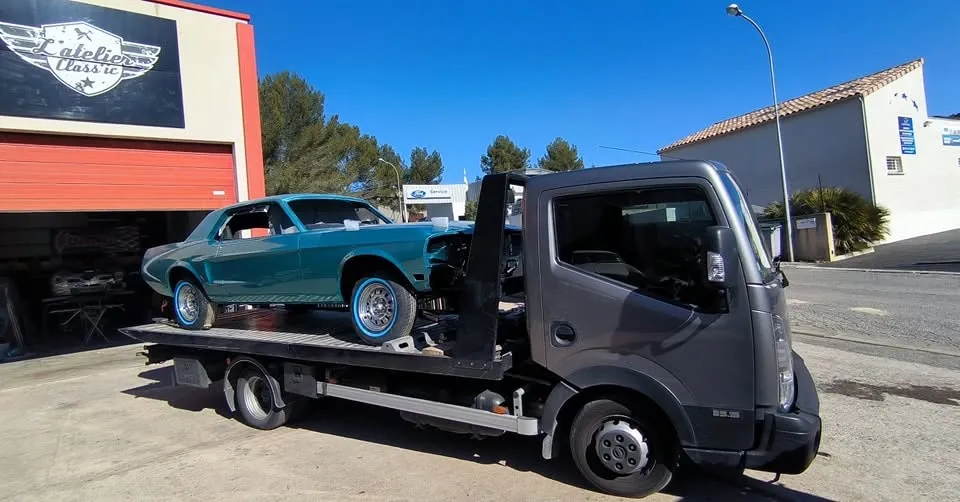 Mustang turquoise partiellement démontée sur un camion plateau devant L'Atelier Class'ic